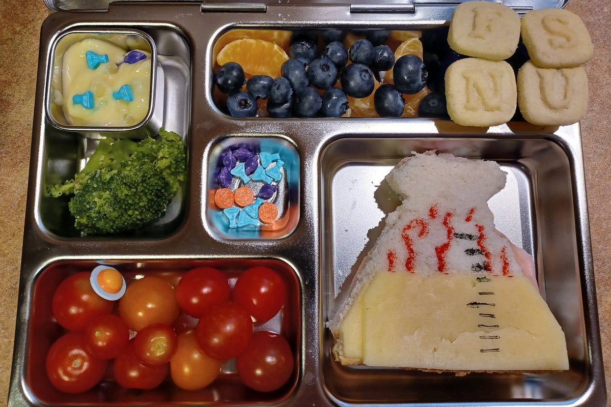 A stainless steel lunchbox filled with a colourful, creative school lunch. The tray includes a sandwich that looks like a steaming beaker. In the other compartments are blueberries, orange slices, broccoli, cherry tomatoes, and letter cookies with the letters F, S, N, U.