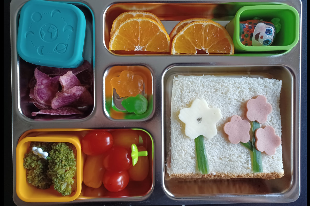 A stainless steel bento lunch box with a spring-time theme. The lunch box contains orange slices, purple chips, broccoli, tomatoes, gummy candies, a ladybug chocolate, and a white sandwich with ham and cheese arranged like flowers.