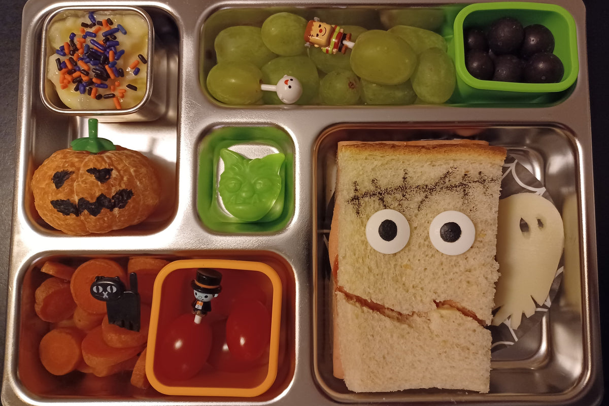 A Halloween-themed lunch in a stainless steel bento box, featuring a sandwich decorated like a Frankenstein face, an orange made to look like a pumpkin, grapes, carrots, cherry tomatoes, a small candy, and pudding with orange, black, and purple sprinkles.