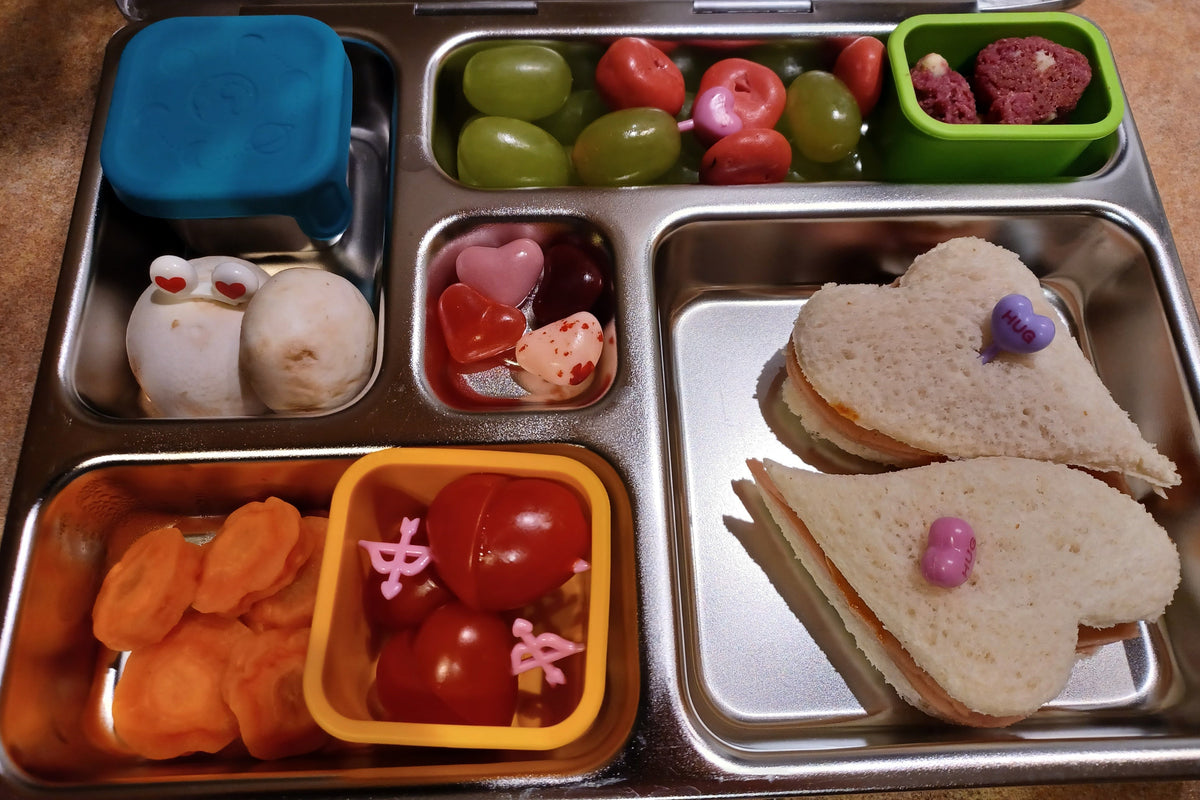 A stainless steel bento lunch box filled with Valentine's themed food: heart-shaped sandwiches, grapes, cherry tomatoes, carrot slices, and mushrooms, and candies, with Valentine's themed food picks.