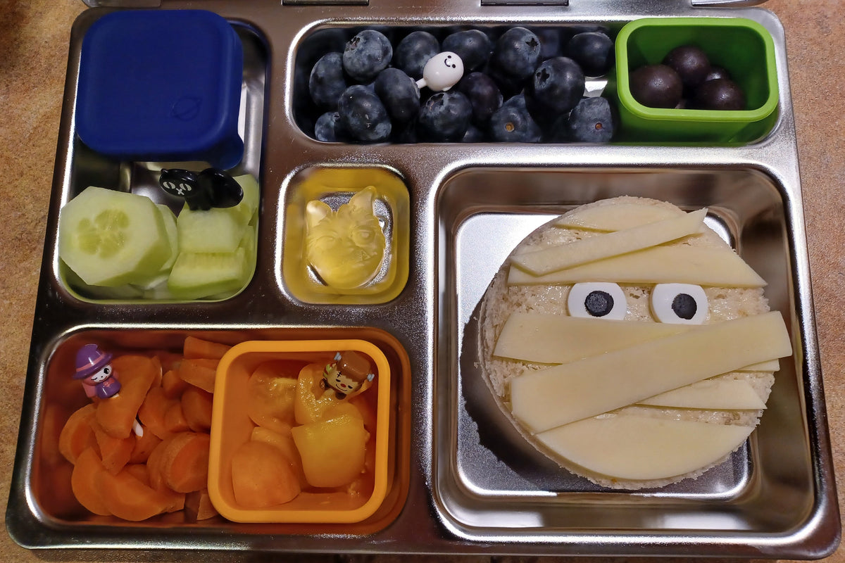 A stainless steel bento lunch box filled with Halloween-themed snacks. The main compartment contains a sandwich styled as a mummy with candy eyes and cheese bandages.