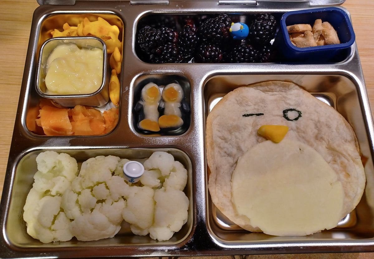 A stainless steel lunchbox filled with a tortilla shaped like a bird face with cheese. In the other compartments are cauliflower, tapioca pudding, blackberries, smoked salmon, penguin crackers, and two penguin gummy candies.