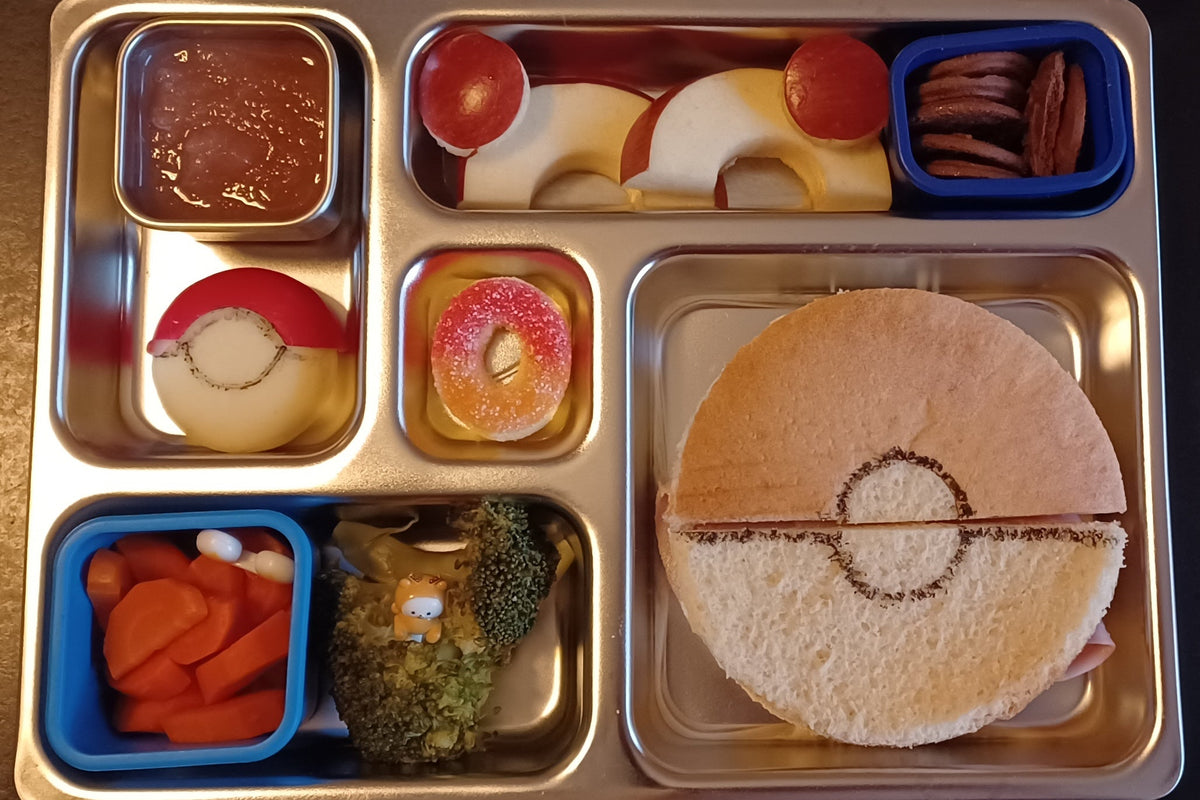 A children's stainless steel bento lunch box filled with themed Pokémon-inspired foods: a sandwich with a Pokéball design on the bread, a circular candy, a Pokéball-shaped cheese, broccoli and carrot slices, and apples cut into Pokéballs.