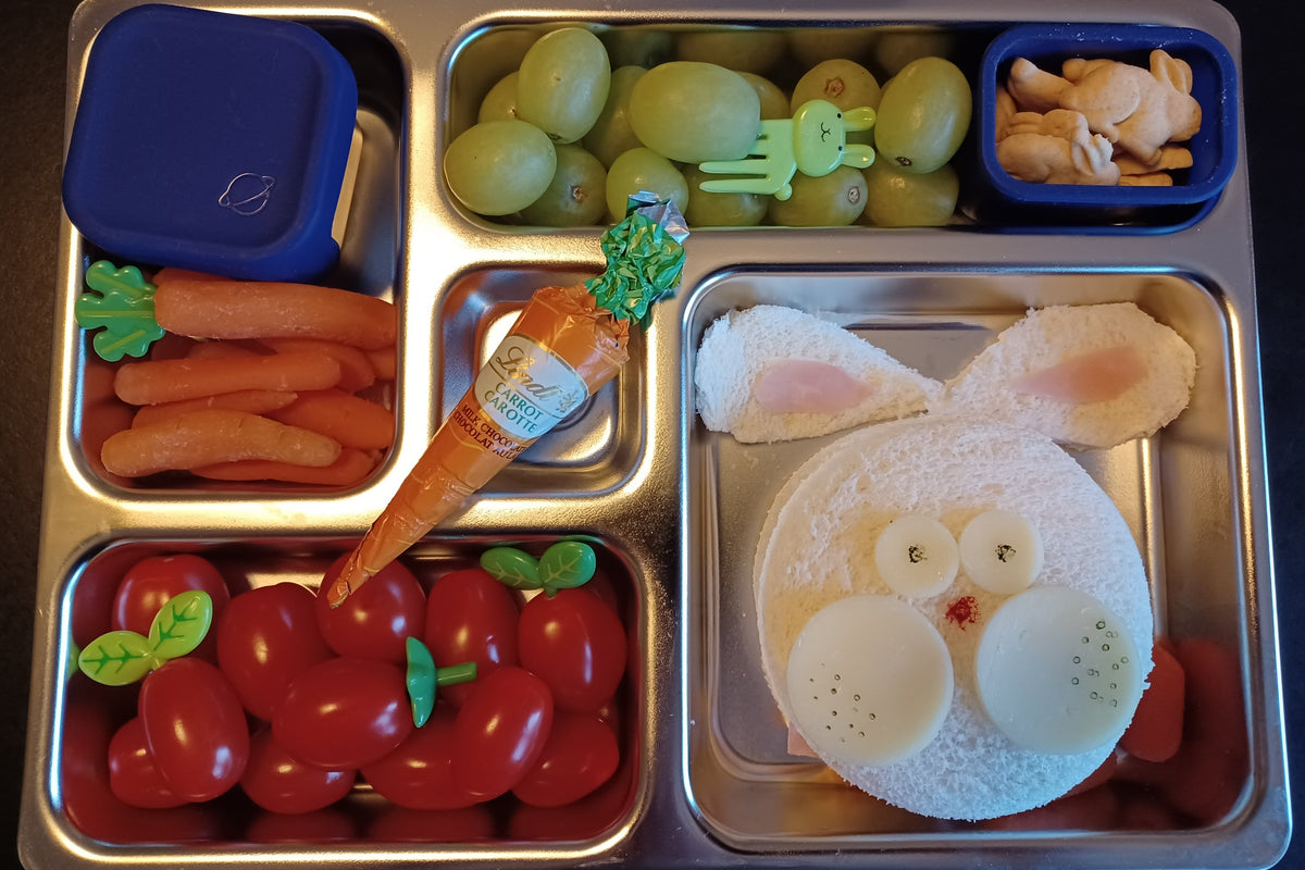 A stainless steel lunch box with multiple compartments containing a rabbit-shaped sandwich, green grapes, baby carrots, cherry tomatoes, bunny cookies, and a chocolate shaped like a carrot.