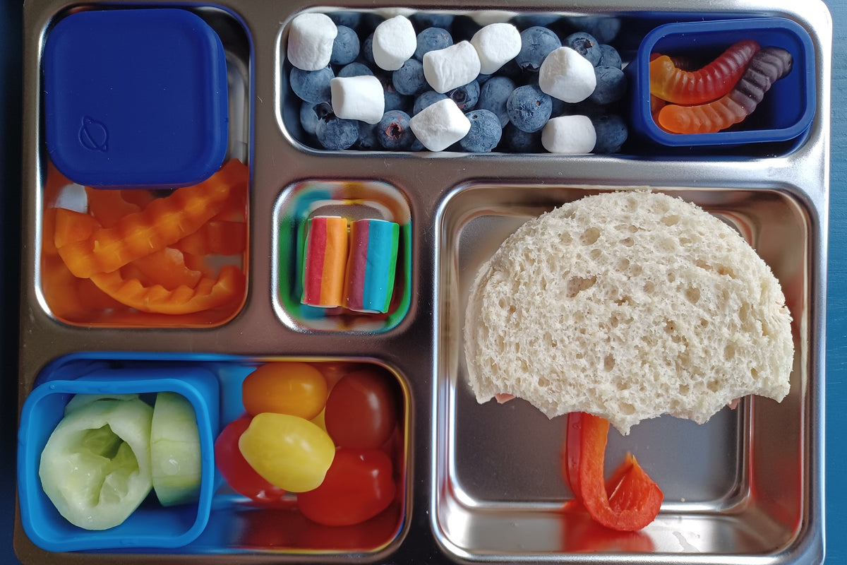 A stainless steel lunch box with multiple compartments containing an umbrella-shaped sandwich with a red pepper handle, blueberries, mini marshmallows, gummy worms, cucumber, cherry tomatoes, and carrot sticks, and rainbow candies.