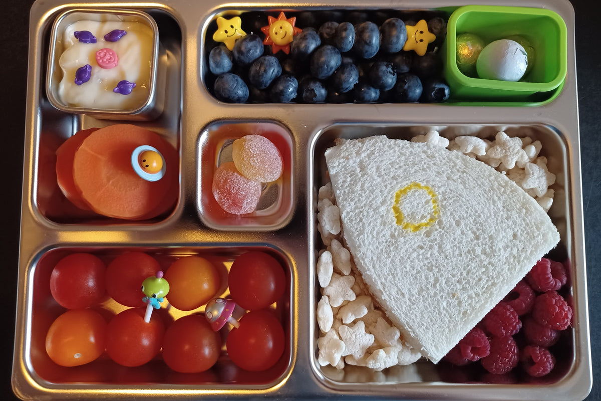 A stainless steel lunchbox filled with a rocket-shaped sandwich, surrounded by star-shaped puffs and raspberries for the flames. In the other compartments are, blueberries with star-shaped plastic food picks, cherry tomatoes with alien food picks, sliced carrots, chocolate spheres, and a small dessert container filled with pudding and planet-shaped sprinkles.