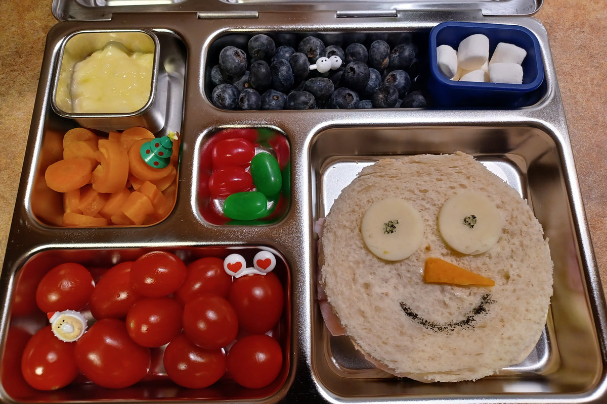 A stainless steel bento lunch box filled with a sandwich shaped like a smiling snowman, blueberries, cherry tomatoes, sliced carrots, pudding, and green and red jellybeans.